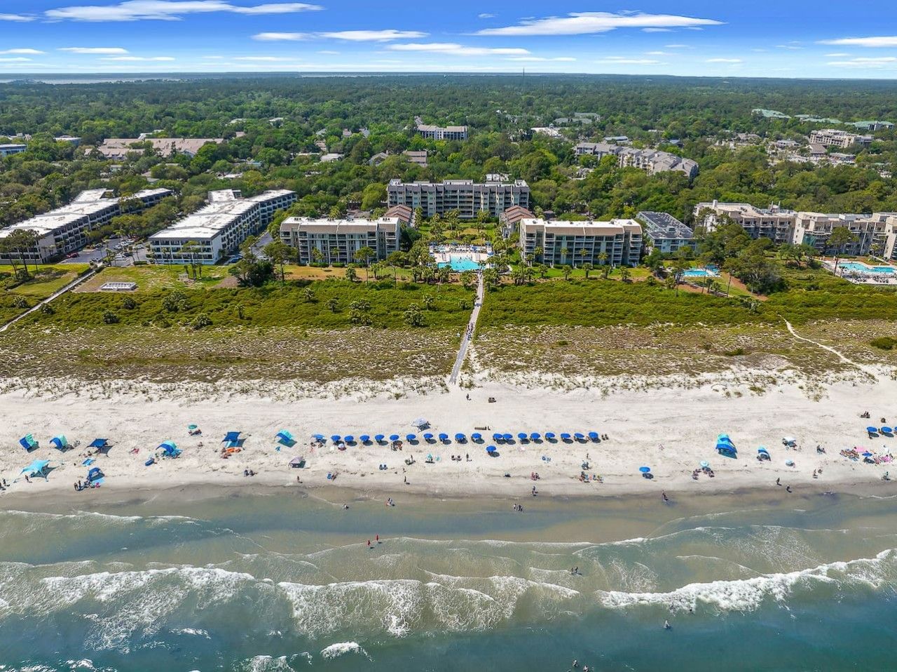 Vue aérienne d’un hôtel situé sur la côte, composé de plusieurs bâtiments, avec une plage de sable bordée de parasols et une vue dégagée sur l’océan.