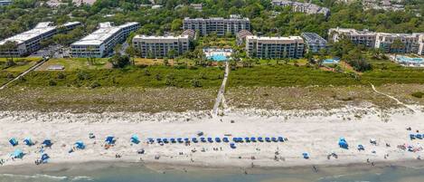 An aerial view of a coastal hotel with multiple buildings, a sandy beach with umbrellas, and a clear view of the ocean.