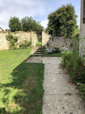 Ein gepflasterter Weg führt zu einer Steinmauer mit einer Tür, umgeben von Grünpflanzen und Blumen.