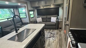 A compact RV kitchen with a central island, built-in cabinets, a stove, and a seating area with a sofa and window views.