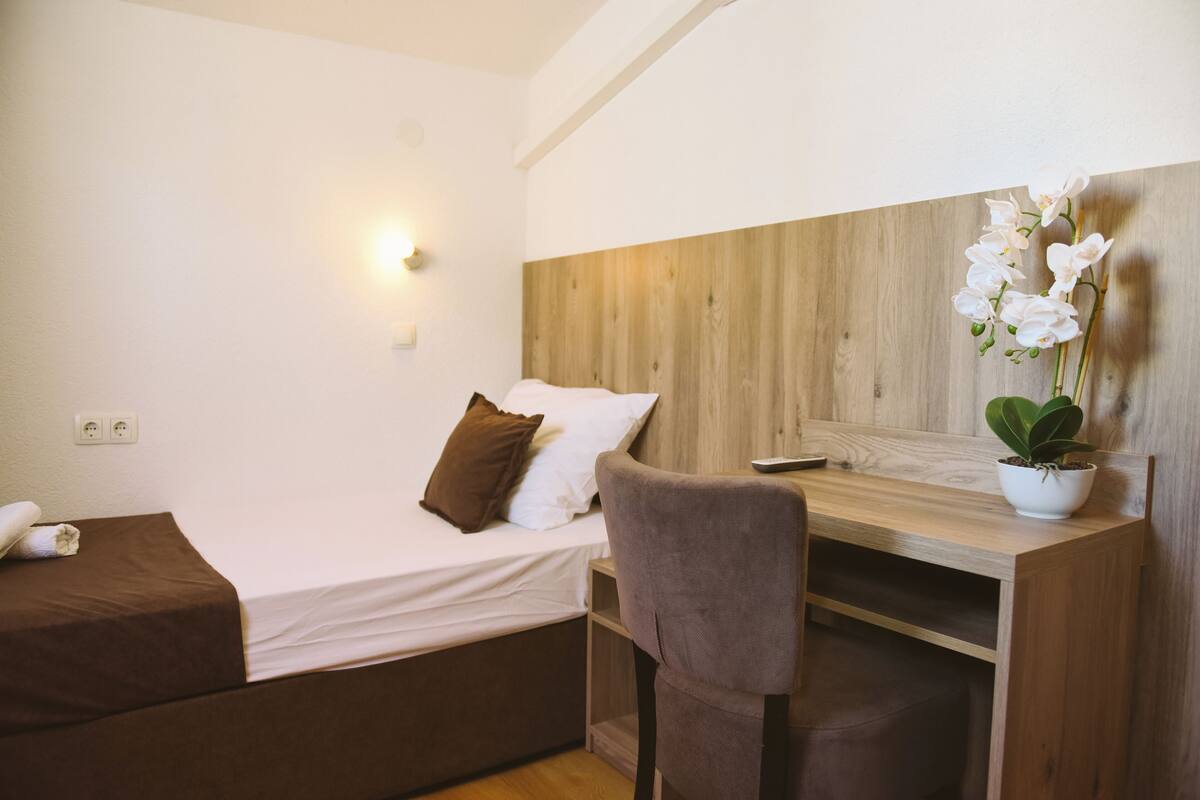 A small, modern room with a single bed, a desk, and a chair. There is a wall-mounted light above the bed and a potted plant on the desk.