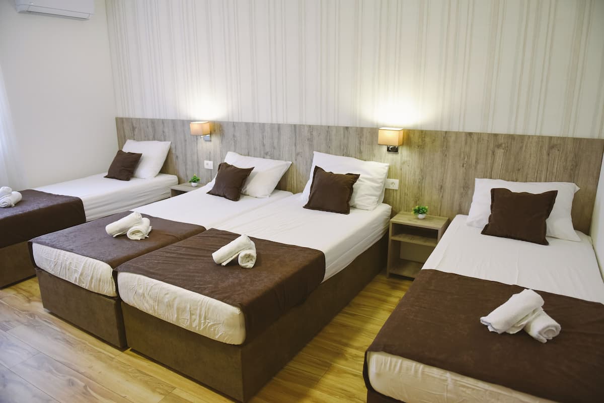 A hotel room with four beds arranged in a row, each with a brown headboard and white bedding. There are two bedside tables with lamps and a small plant on one. The room has wooden flooring and a striped wallpaper design on the walls.