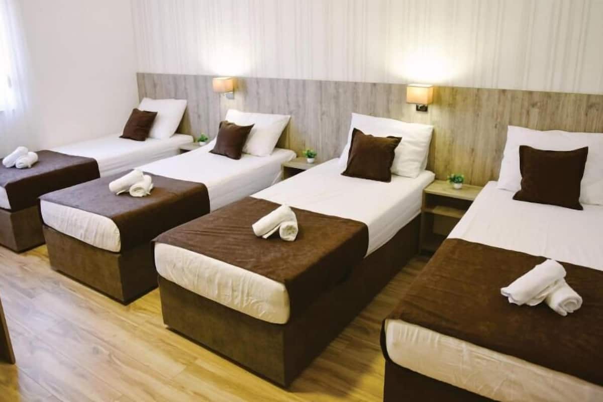 A hotel room with four single beds, each with a brown headboard, white linens, and a rolled towel on the foot of the bed.