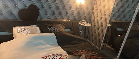 A bedroom with a tufted headboard, a heart-shaped pillow, and a bedspread with a heart design and the text "YOU & ME".