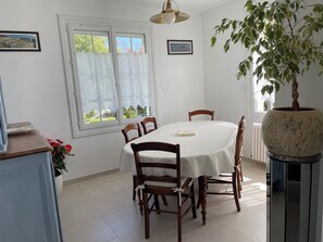 A dining area with a round table, wooden chairs, a potted plant, and a window with curtains. - 🌊 Near the sea, private courtyard and large enclosed garden 🌲 2 to 5 persons (Reville)