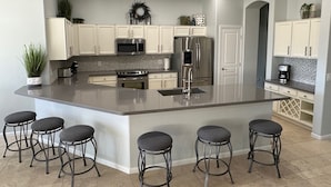 A modern kitchen with white cabinets, stainless steel appliances, a large island with bar stools, and a decorative wreath on the wall. - Water/Golf Course Backyard | Heated Pool Included in Price | Modern 3 bedroom (Chandler)