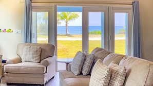 Ein Wohnzimmer mit einem beigen Sofa und einem Sessel, einem kleinen Tisch und Blick auf Strand und Palme durch große Glasüren.