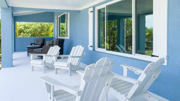 Un espacio de estar al aire libre con techo, sillas tipo Adirondack y una mesa, frente a una pared azul con ventanas amplias.