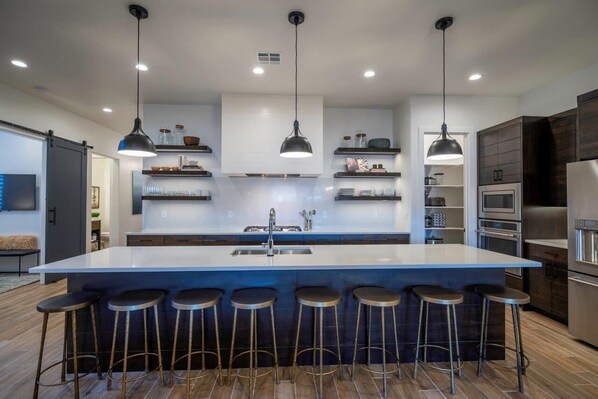 A modern kitchen with a large island, bar stools, pendant lights, and stainless steel appliances.