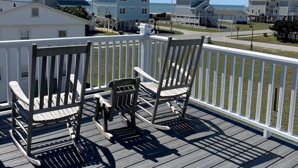 Een houten terras met twee schommelstoelen, een witte balustrade en uitzicht op strandhuizen en de oceaan.