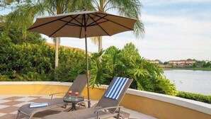 A sun lounge with two chairs and a table, an umbrella, and a view of a body of water and greenery.