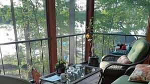 Un espacio al aire libre techado con mobiliario de mimbre, una mesa con cubierta de vidrio y vistas a árboles a través de amplios ventanales.
