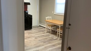 Una habitación con suelo de madera, una mesa de comedor con sillas, una ventana con persianas y una zona de cocina con estufa y armarios.