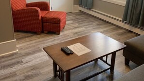 Un salón con suelo de madera, una mesa de centro marrón con un control remoto, una silla roja con estampado, un sofá gris, un televisor de pantalla plana montado en la pared y una ventana con cortinas.