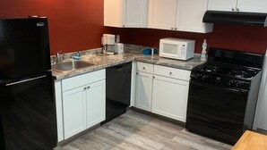 Una cocina compacta con armarios blancos, un refrigerador negro, un microondas, una estufa y una mesa de madera.