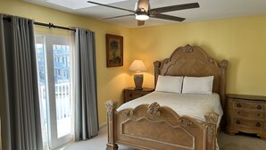 A bedroom with a wooden bed, a ceiling fan, a nightstand with a lamp, and a view through a window with curtains.