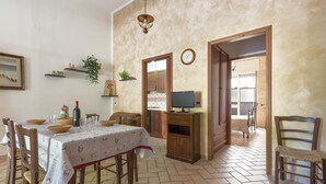 A dining area with a table set for four, wooden chairs, a television on a stand, and a kitchen visible through an open door. - 2344 Le Due Torri - Torre Piccola by Barbarhouse (Melendugno)