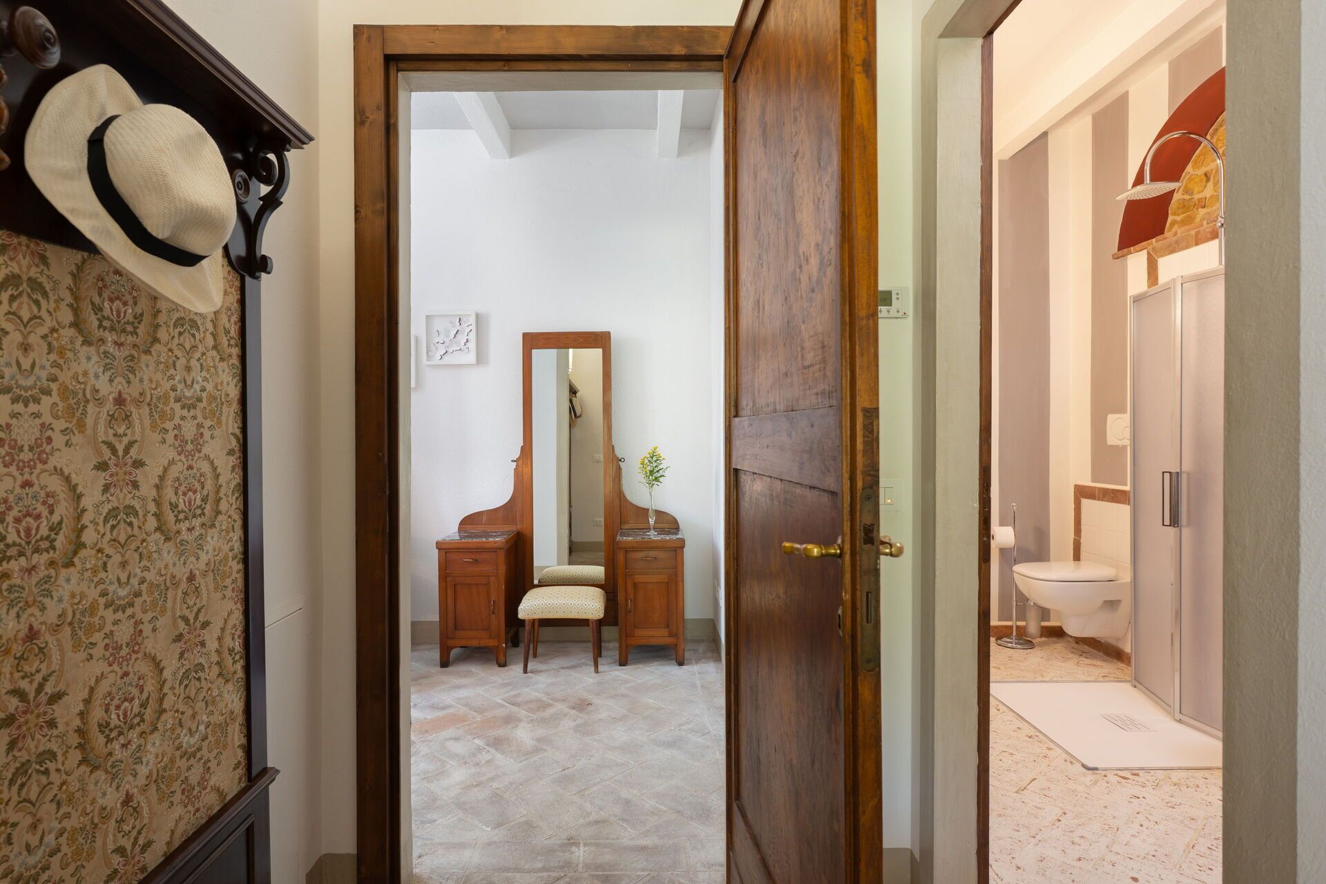 Un couloir mène à une salle de bain équipée d’une douche, d’un meuble-lavabo avec un miroir et d’une porte dotée d’une poignée en laiton.