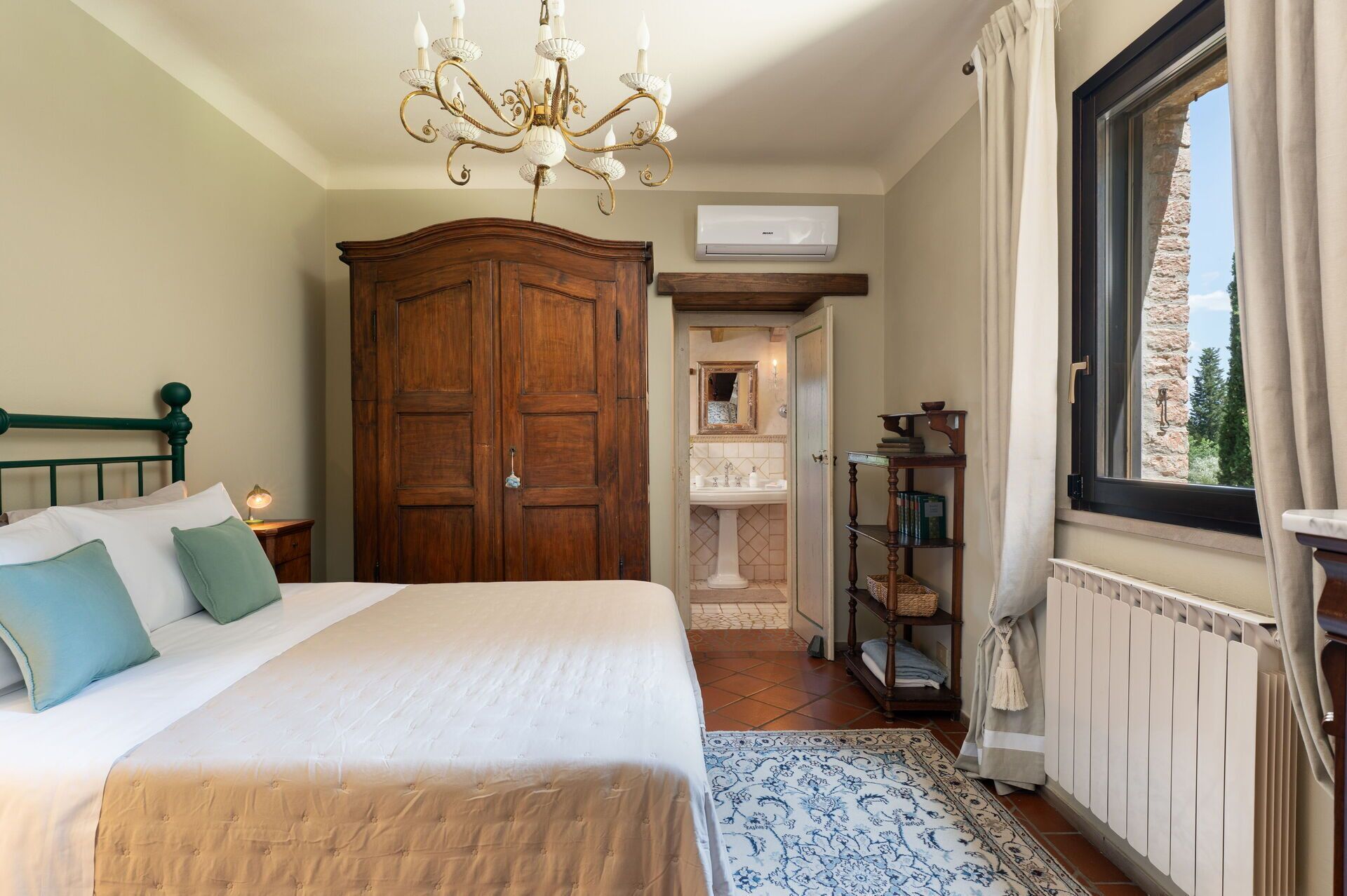 Une chambre avec un grand lit, une armoire en bois, un lustre, une fenêtre avec des rideaux, un radiateur et une salle de bain visible à travers une porte ouverte.
