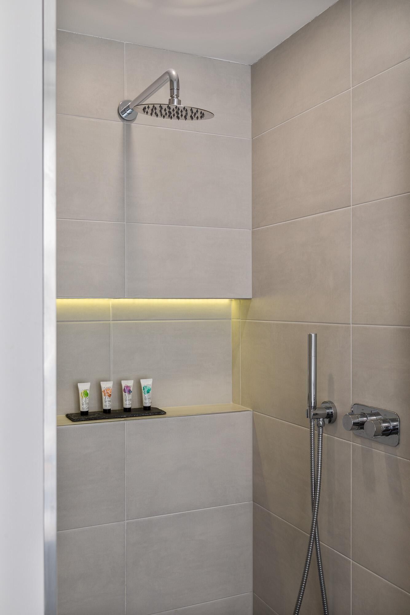 Une douche moderne dotée d’un pommeau de douche à main et d’un robinet mural. On trouve trois articles de toilette sur une étagère située en dessous.