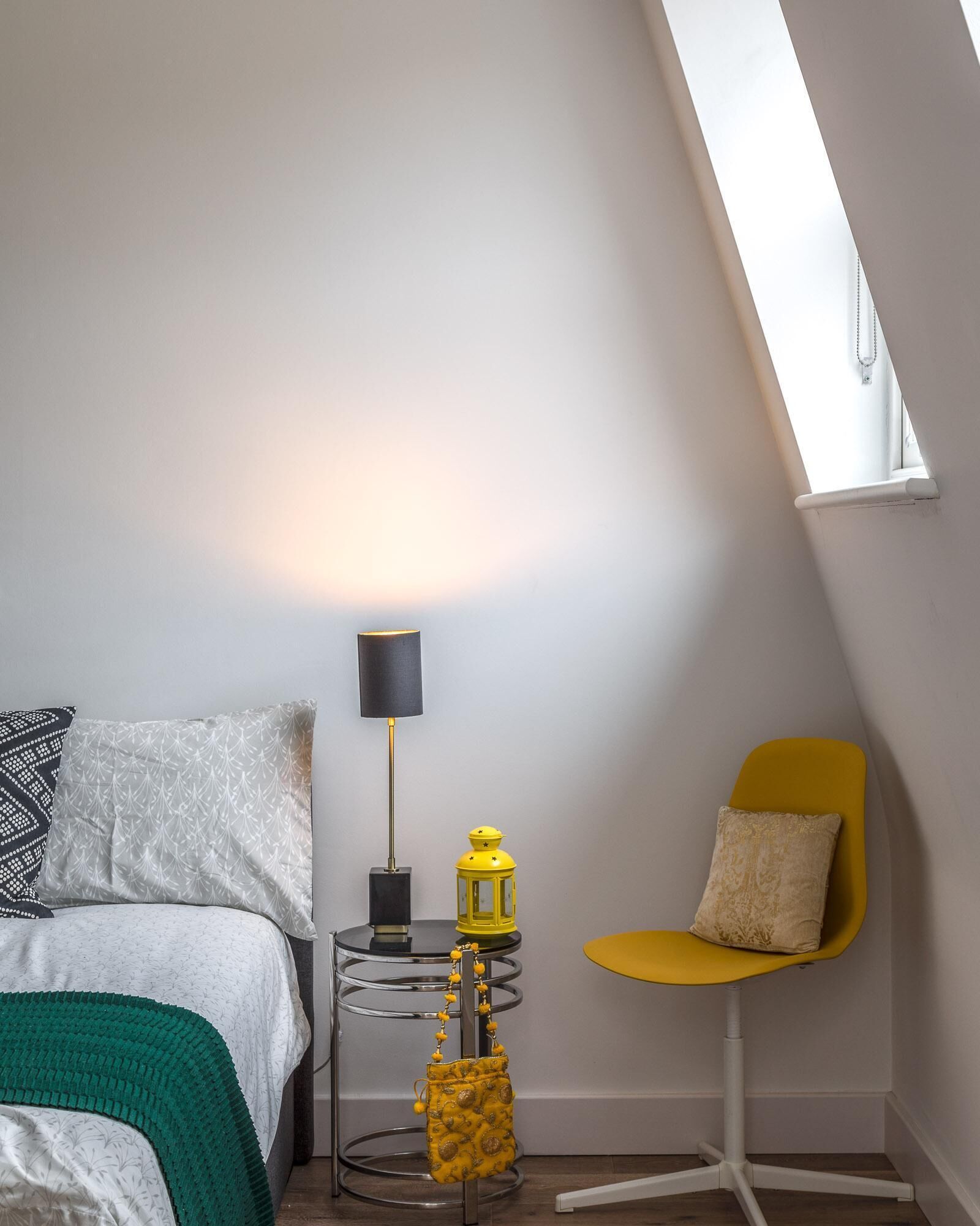 A bedroom with a bed, a bedside table with a lamp, a small table with a lantern, a chair, and a skylight.