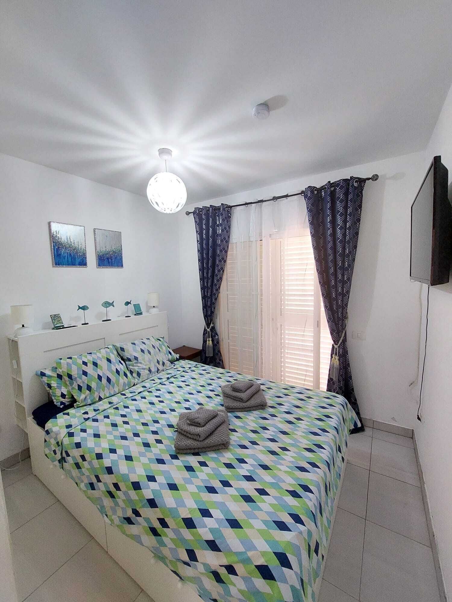 Une chambre avec une couvre-lit à carreaux, une tête de lit blanche, deux décorations murales représentant des poissons, un luminaire au plafond et un téléviseur fixé au mur.