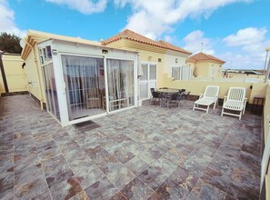 Ein überdachter Patio-Bereich mit Esstisch und Stühlen, einer Schiebetür aus Glas und einem gefliesten Boden.