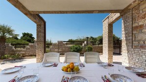 Area pranzo all'aperto con tavolo apparecchiato per quattro persone, circondata da muri in mattoni e sedie bianche.