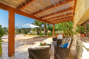 An outdoor patio with wicker furniture, a thatched roof, and a view of palm trees and a white umbrella.