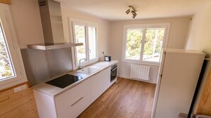 Cocina compacta con muebles blancos, estufa integrada, pileta y refrigerador. Dos ventanas ofrecen luz natural y vistas a la vegetación exterior.
