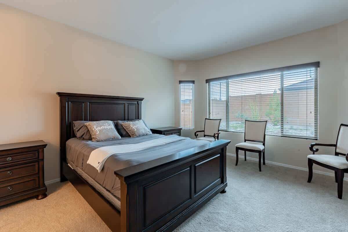 A bedroom with a large bed, two chairs, a nightstand, and a window with blinds.