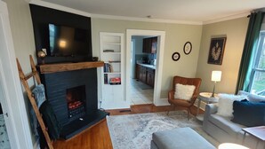 Sala de estar con chimenea, un sofá, una mesa de centro y una lámpara. Se ve una cocina a través de un vano abierto.