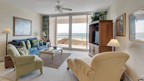 Een woonkamer met een bank, fauteuils, een salontafel en een televisie. Er is een balkon met uitzicht op zee.