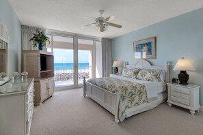 Une chambre à coucher avec un lit, deux tables de chevet, un ventilateur de plafond, une grande fenêtre donnant sur la plage et un tableau accroché au mur.