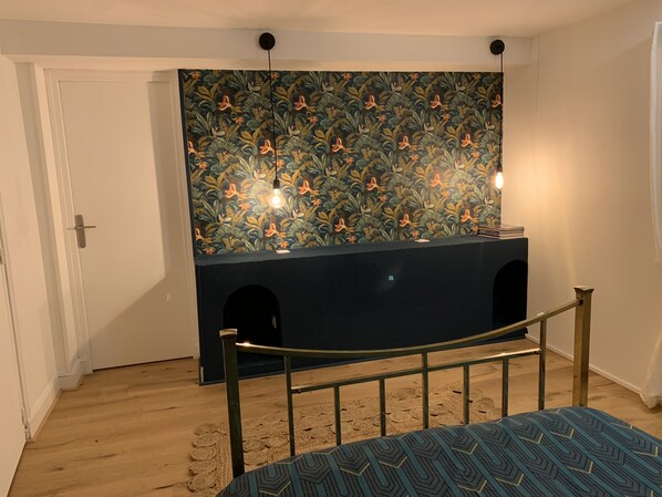 A bedroom with a bed, a dark-colored dresser with two light fixtures, a door, and a patterned wallpaper.