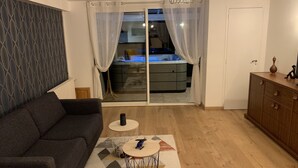 Ett vardagsrum med en mörk soffa, ett litet runt soffbord och en mönstrad matta. En skjutdörr i glas leder till ett köksområde, och genom glaset syns en säng.