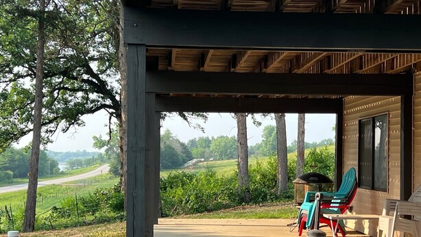 A covered outdoor seating area with chairs and a table, overlooking a rural landscape with trees and a road. - Beautiful house on Lake Rathbun with in-ground pool & 50-amp camper hookup (Plano)