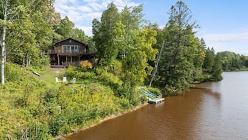 A cabin with a deck and a boat dock, surrounded by dense forest and a river.
