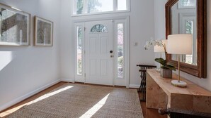 Un pasillo luminoso con una puerta blanca, un espejo grande y una consola de madera con una lámpara.