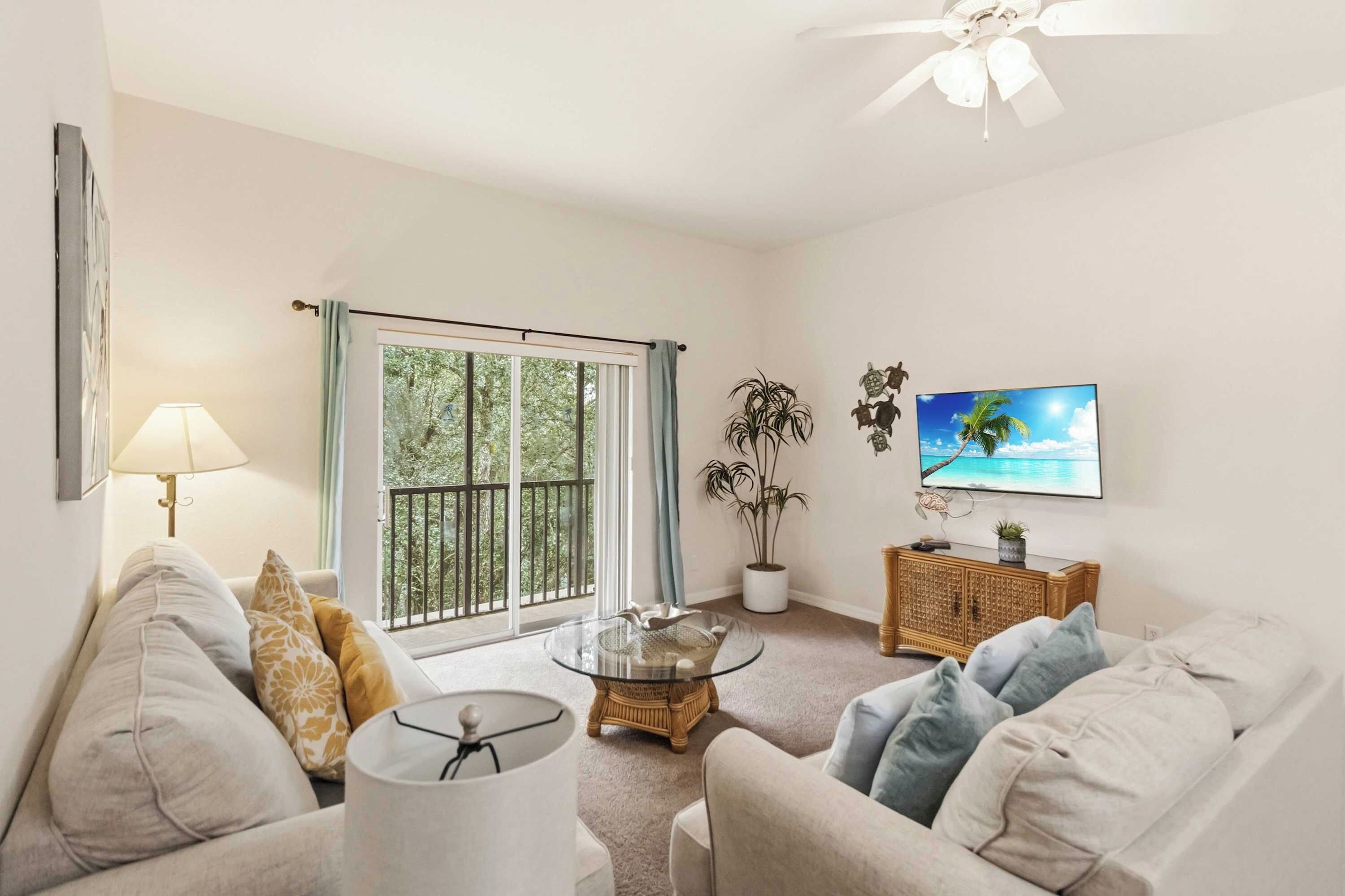 A living room with a sofa, a coffee table, a TV, and a balcony with a view of trees.