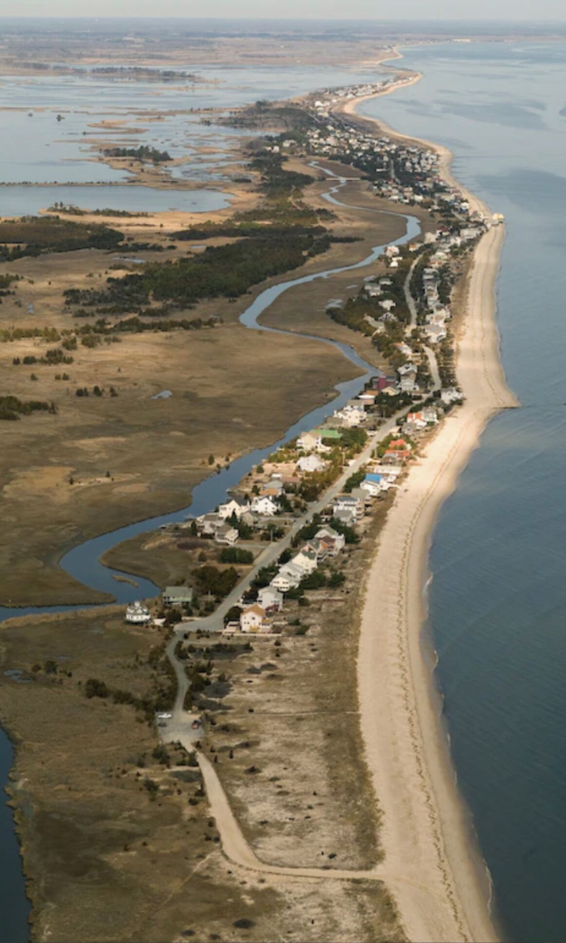 An aerial view of a coastal town with a long stretch of beach, a river running parallel to the shore, and a series of houses along the coastline.