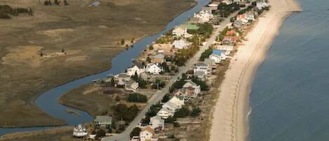 An aerial view of a coastal town with a long stretch of beach, a river running parallel to the shore, and a series of houses along the coastline.