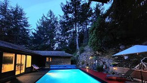 Une piscine dotée d’une terrasse en bois et de chaises longues, entourée d’un mur de pierre et d’arbres.