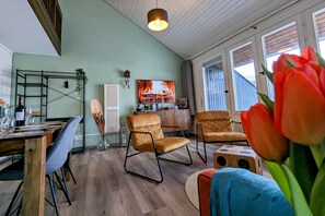 A modern living room with a wooden dining table, two cushioned chairs, a television, and a large window with a view of the outside.