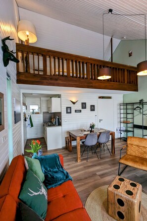 A spacious living area with a high ceiling, wooden ceiling beams, and a dining area with a kitchen in the background.