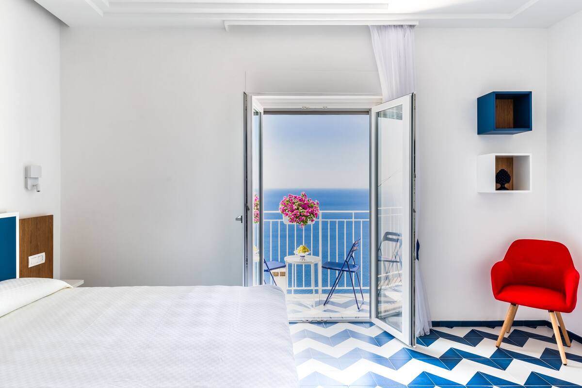 A hotel room with a bed, a red chair, a balcony with a view of the sea, and a small table with flowers.