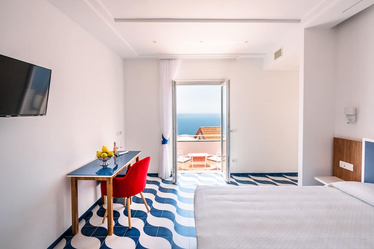 A hotel room with a bed, a desk with a chair, a TV mounted on the wall, and a balcony with a view of the sea.