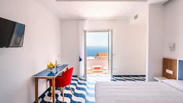 A hotel room with a bed, a desk with a chair, a TV mounted on the wall, and a balcony with a view of the sea.