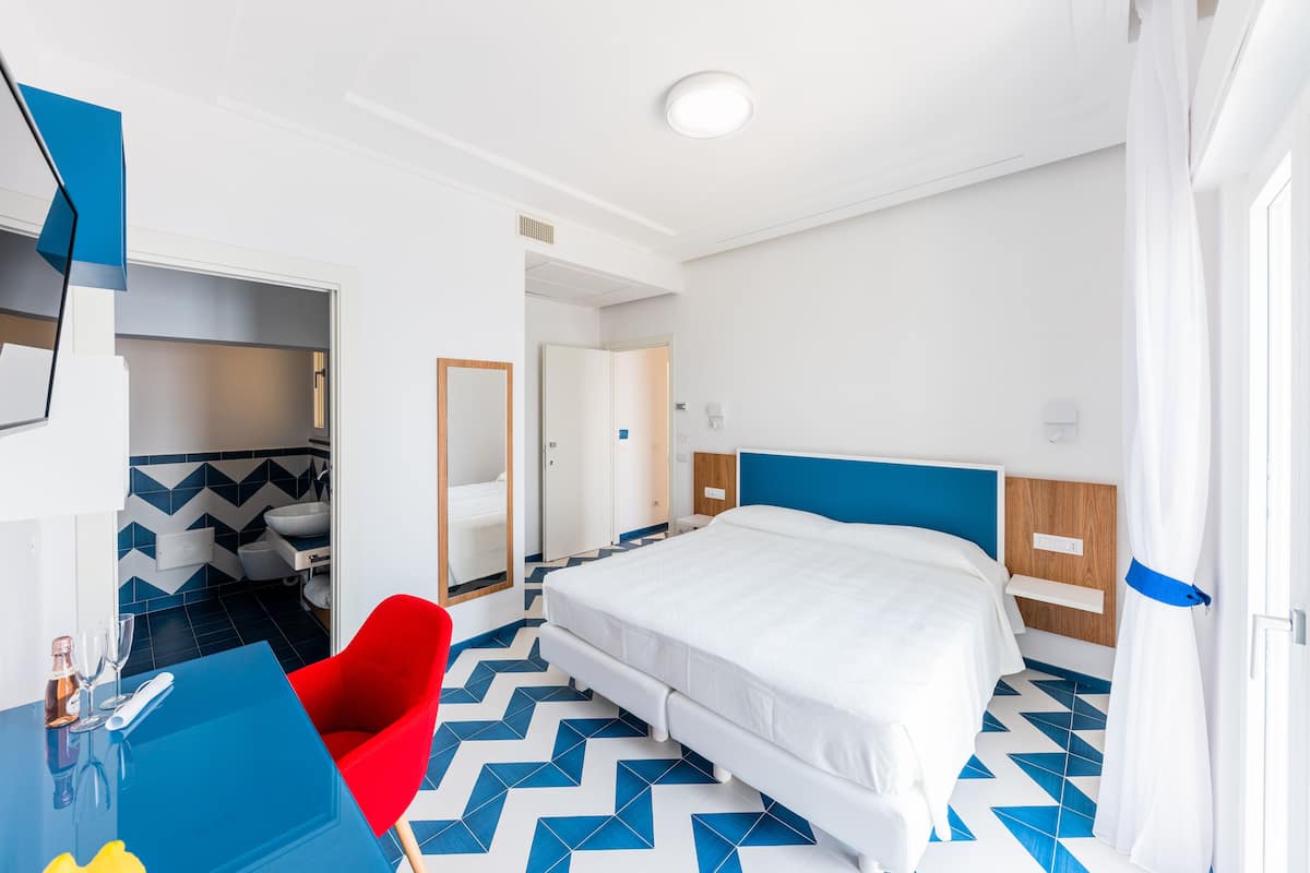 A hotel room with a bed, a red chair, a blue table, and a bathroom visible through an open door.