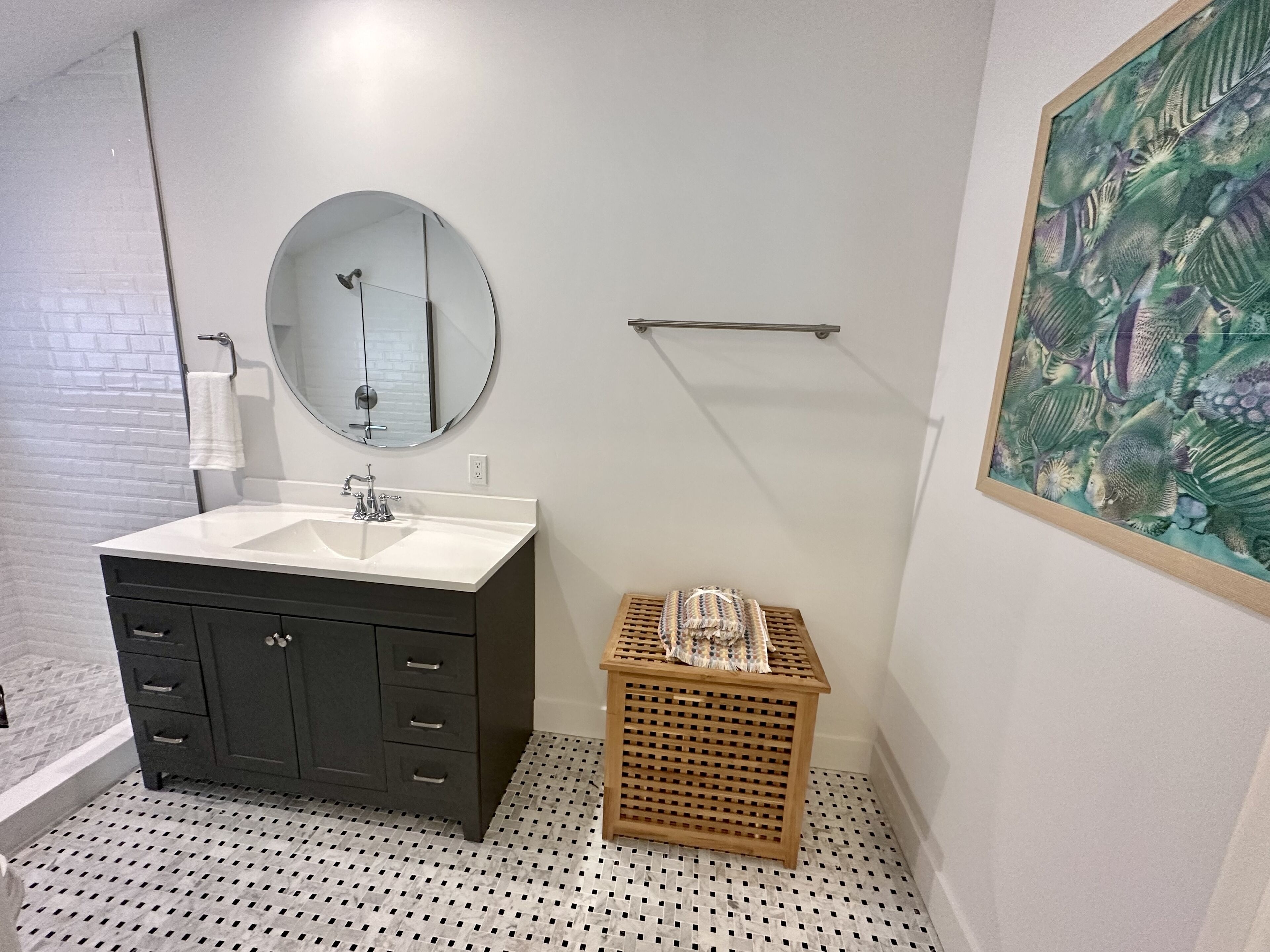 A bathroom with a white sink, dark cabinets, a round mirror, a wooden basket, and a framed fish painting on the wall.